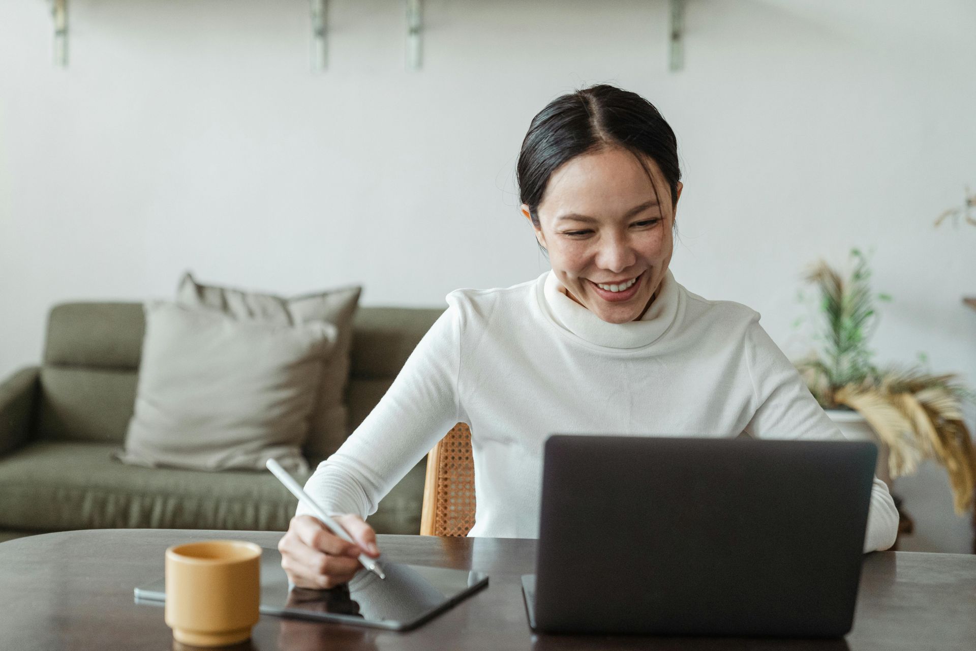 Woman in white turtleneck smiles while using a laptop and tablet, with a cup and couch in the background.