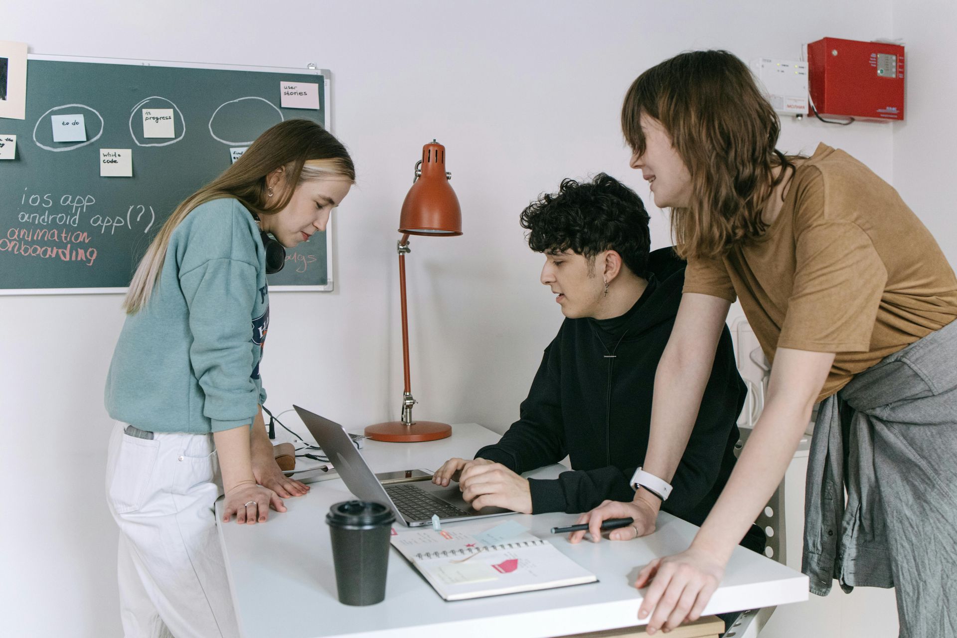 Three people collaborating around a laptop at a desk. One points, and another looks on, near a chalkboard.