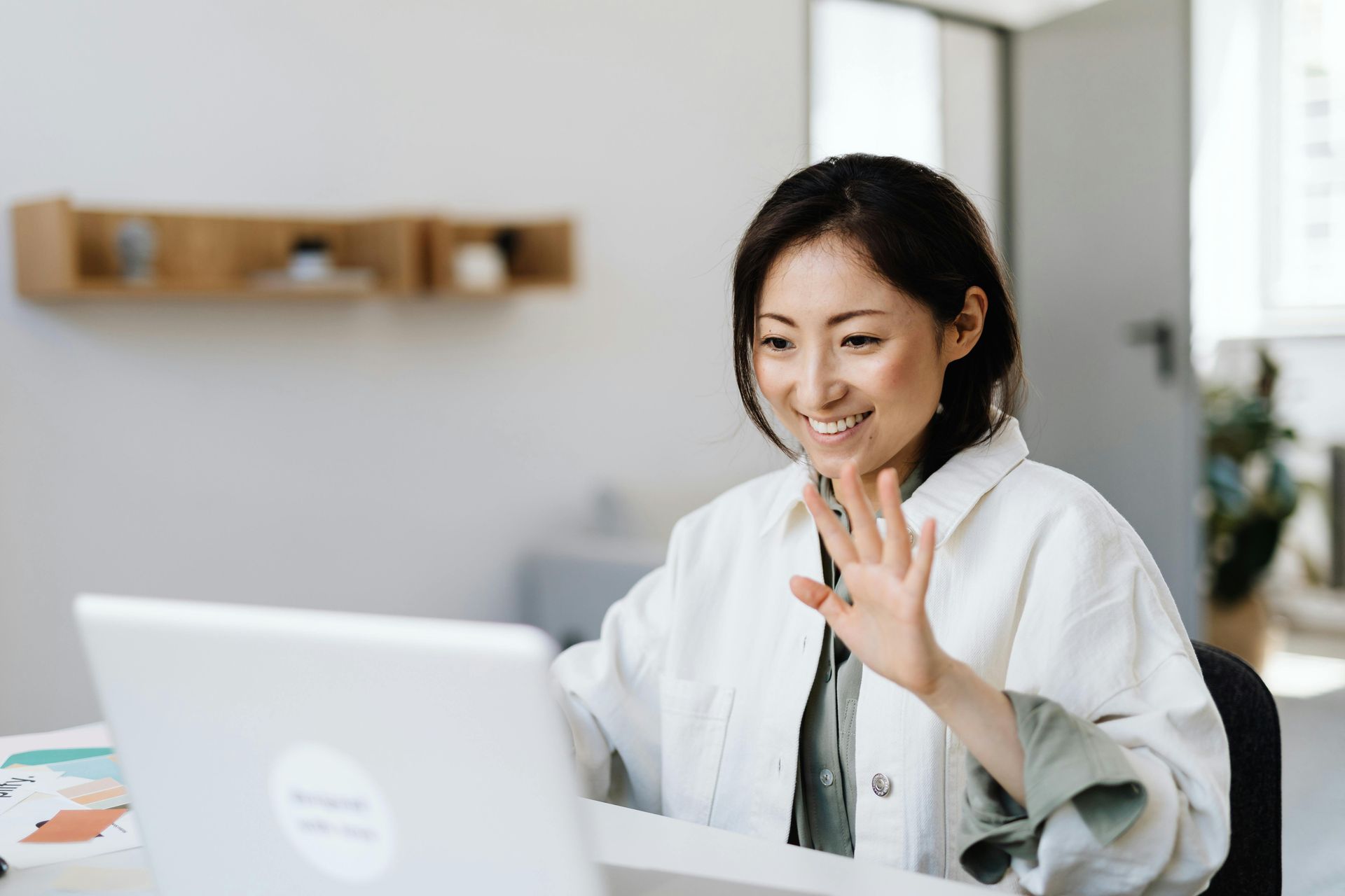 Woman in a white jacket waves at a laptop screen, smiling, in a bright office setting.