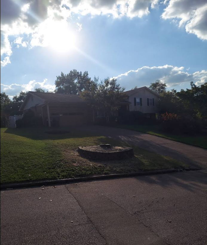 House with carport and driveway, bright sun, clouds, and green lawn.