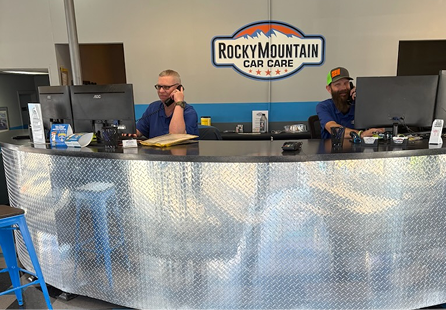 Two men at a Rocky Mountain Car Care counter on phones, using computers, behind a diamond plate desk. | Rocky Mountain Car Care