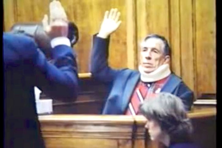 Man in neck brace raising hand in courtroom, taking an oath. A person stands to his left, and a woman sits below.