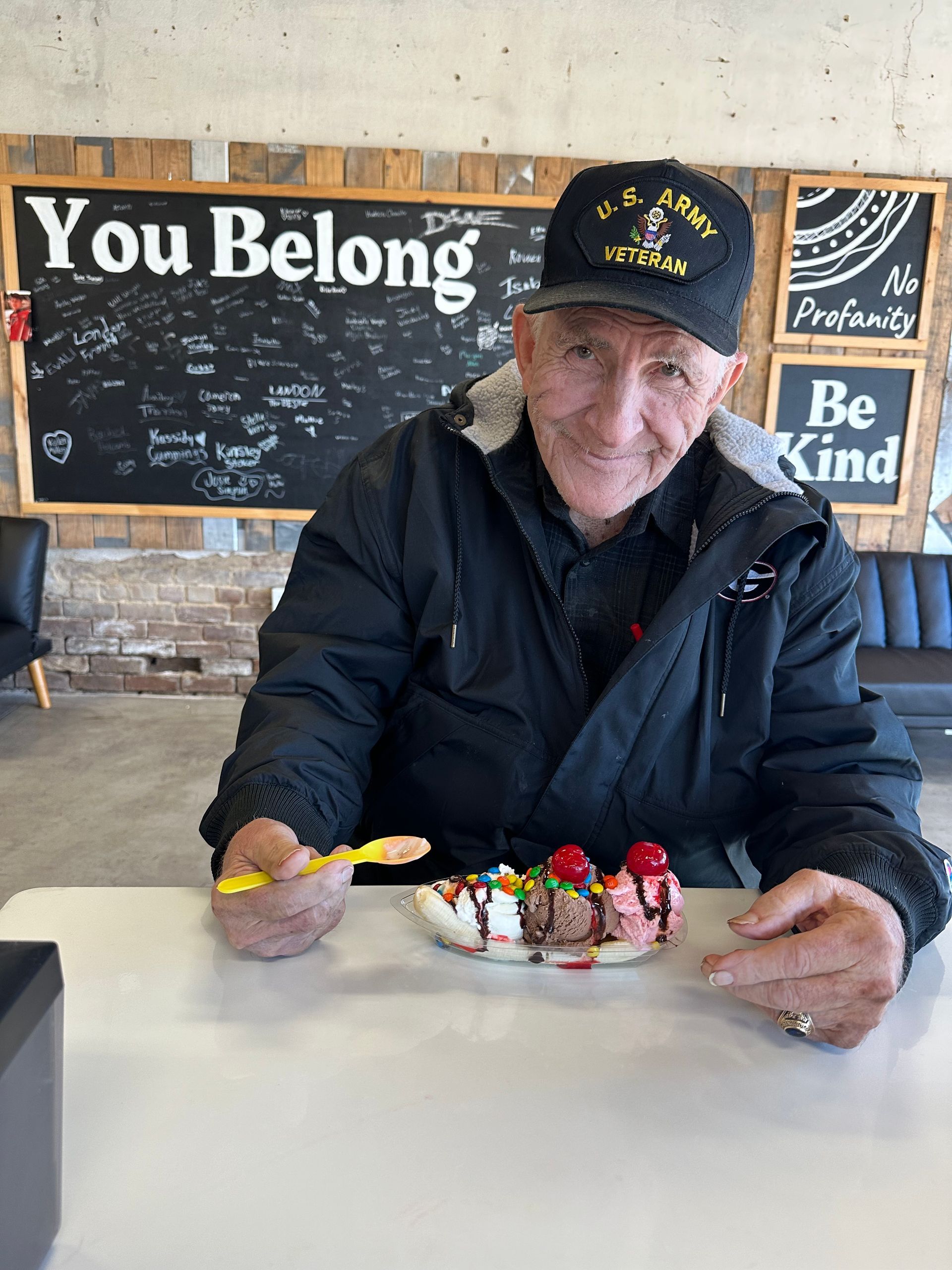 Older man smiles, holding a spoon over a banana split. He's wearing an Army hat and jacket, sitting at a table in a cafe. 