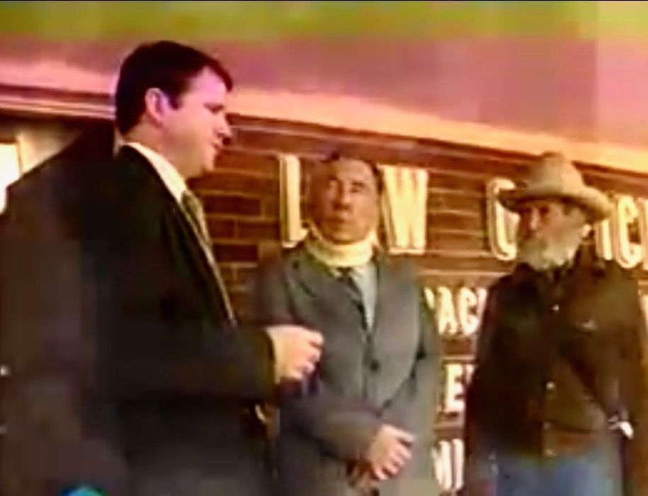 Three men stand outdoors, one in a suit speaking, one in a neck brace, and one in a cowboy hat. They appear in front of a building.