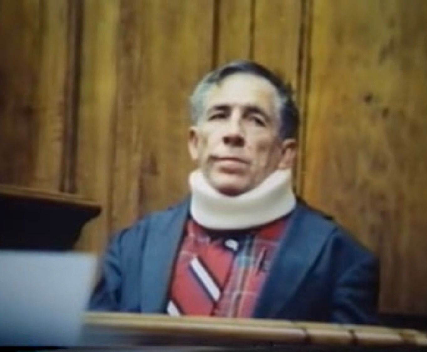 Man in courtroom wearing a neck brace, sitting in front of wooden paneling. He has a serious expression and is wearing a blue blazer and red patterned tie.