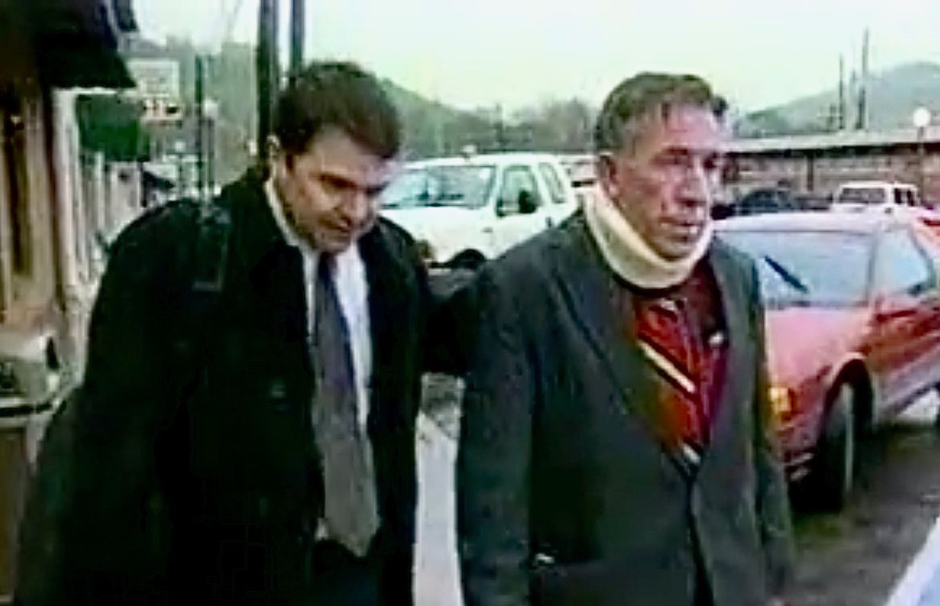 Two men walking outside, one wearing a neck brace and a suit. The other man is in a dark overcoat and holding a briefcase.