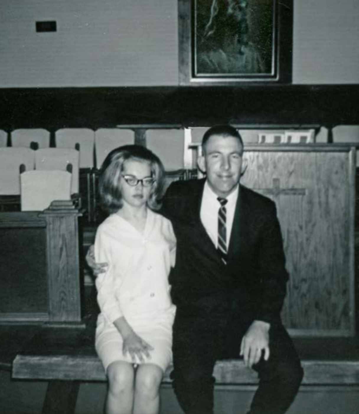 A man and woman sit on a wooden bench. The man wears a suit and tie, and the woman wears a light-colored dress and glasses. They are indoors.