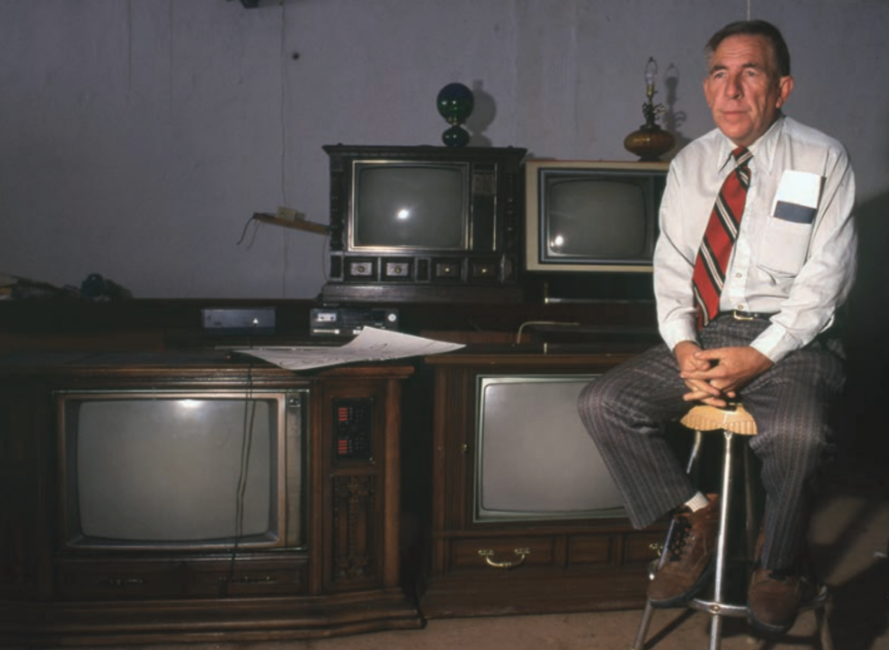 Man in a button-down shirt and tie sits on a stool in front of multiple vintage televisions. Brown wooden cabinet.