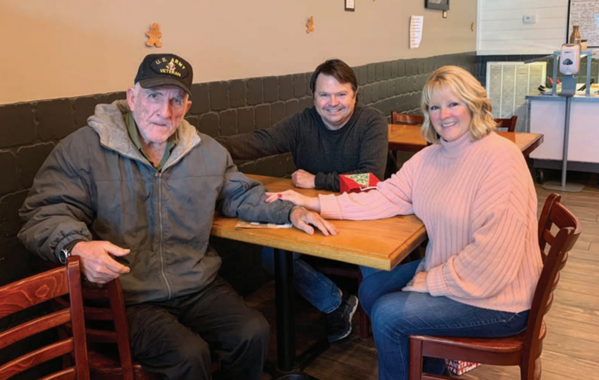 Three people sit at a table in a restaurant. An older man in a jacket and cap, a man in a sweater, and a woman in a pink sweater smile at the camera.