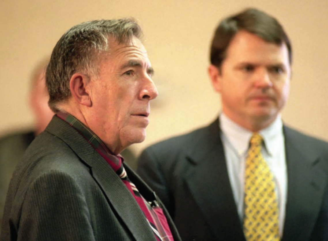 Older man with furrowed brow in a courtroom, wearing a blazer and plaid shirt, next to a man in a suit and tie.