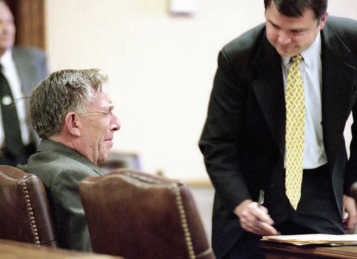 An older man seated in a courtroom appears to be listening to a younger man in a suit who is leaning over a document.