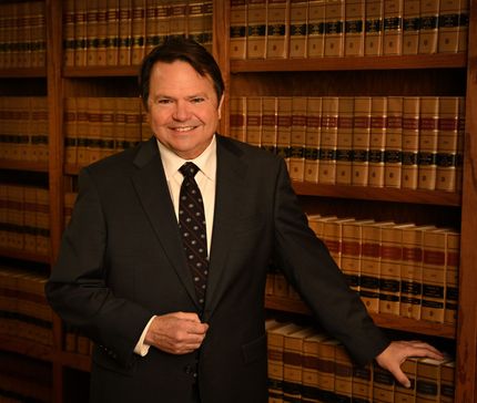 A man in a suit stands in a library, smiling and touching a bookshelf. He has dark hair and is wearing a black tie.