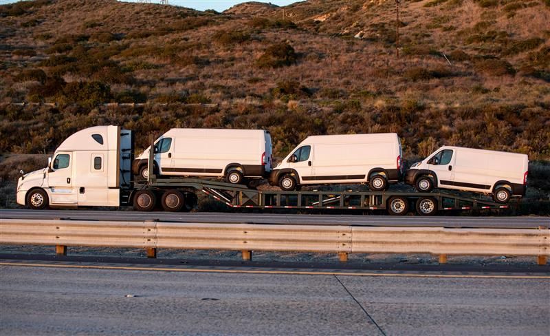White semi-truck hauling three white vans on a trailer, driving on a highway; hills in the background.