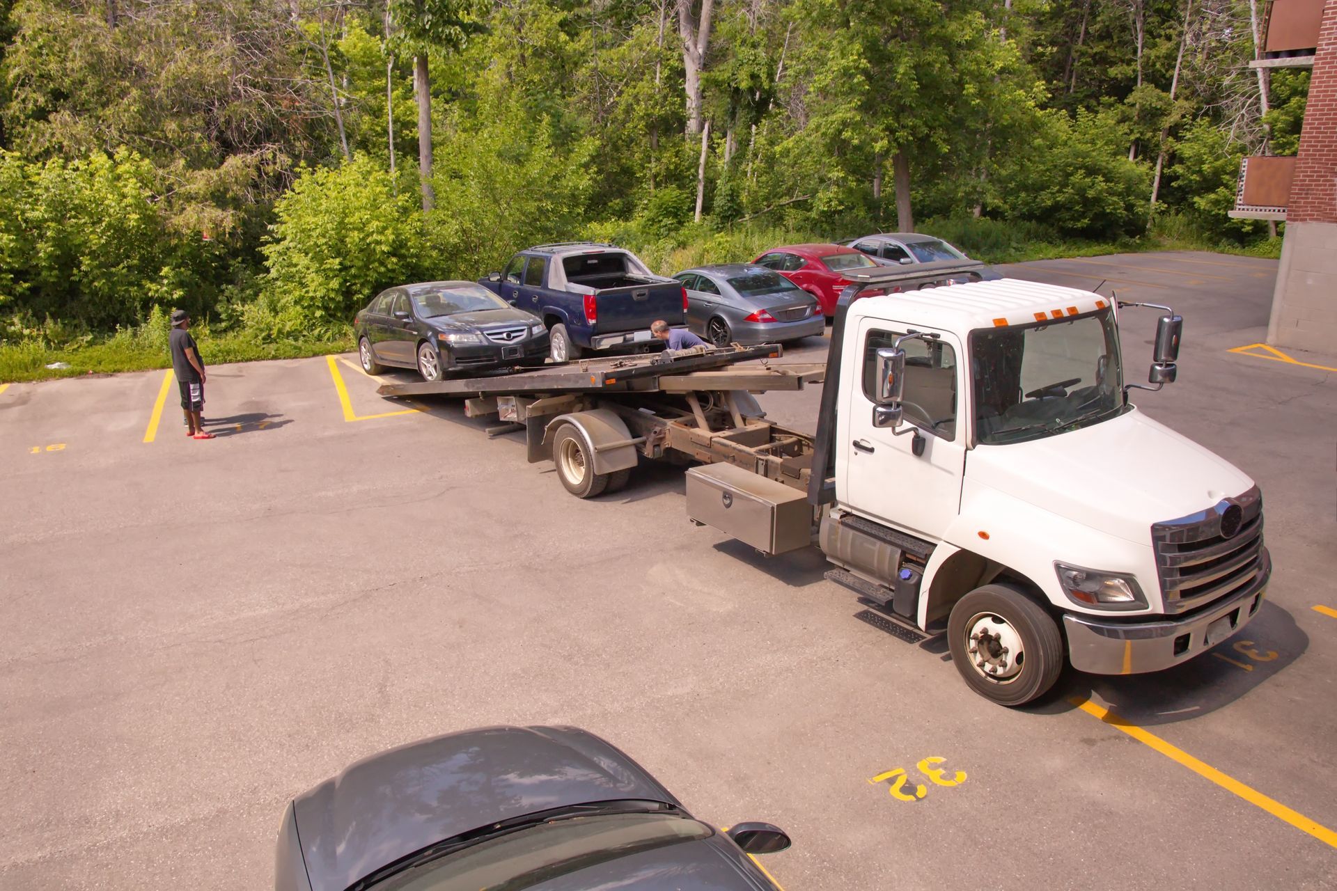 Tow truck with multiple cars parked in an asphalt lot, person in the background, trees and building.