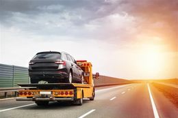 Black car being towed on a highway by a yellow tow truck under a cloudy sky.