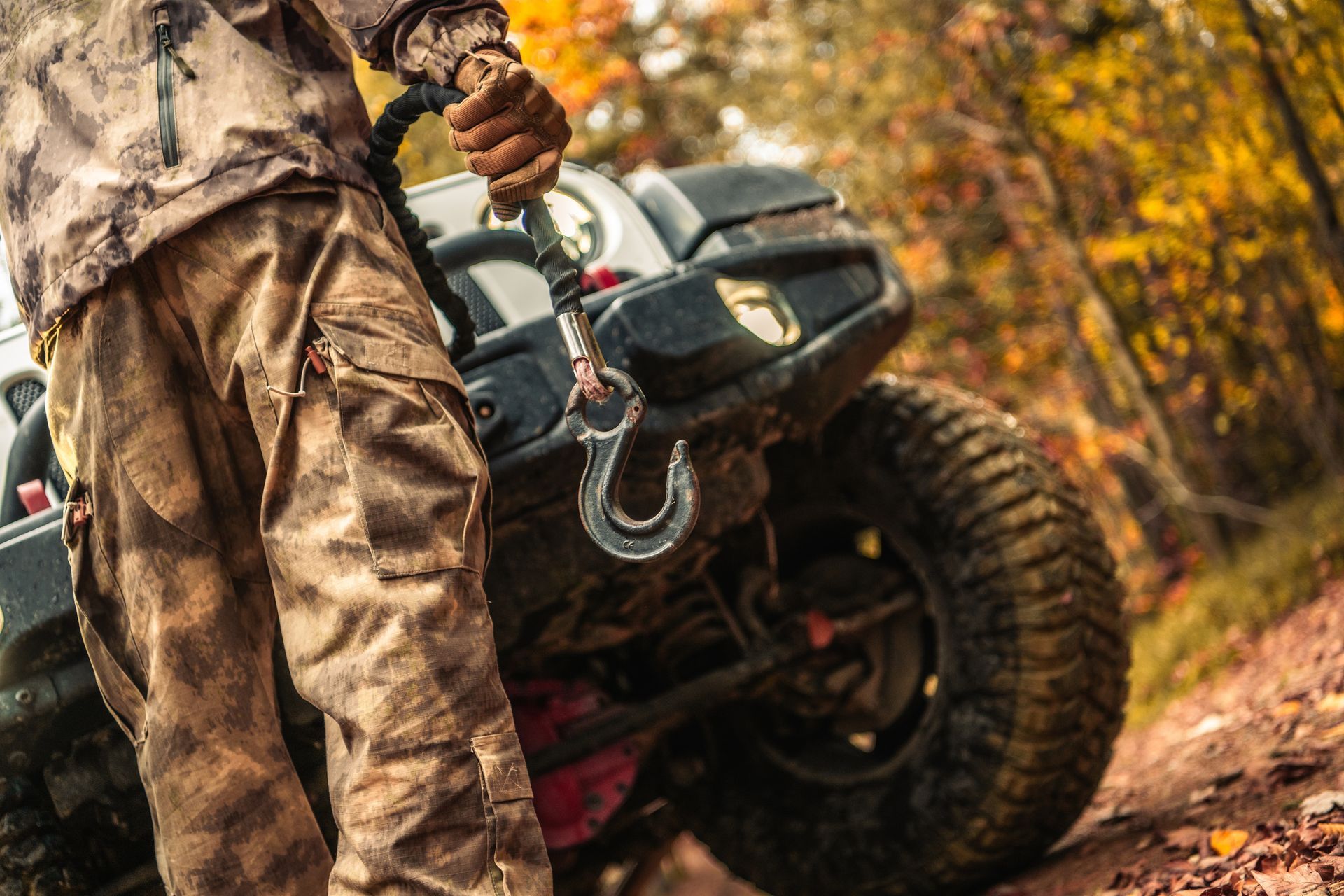 Person in camo gear attaching a hook to a winch on a Jeep, autumn forest background.
