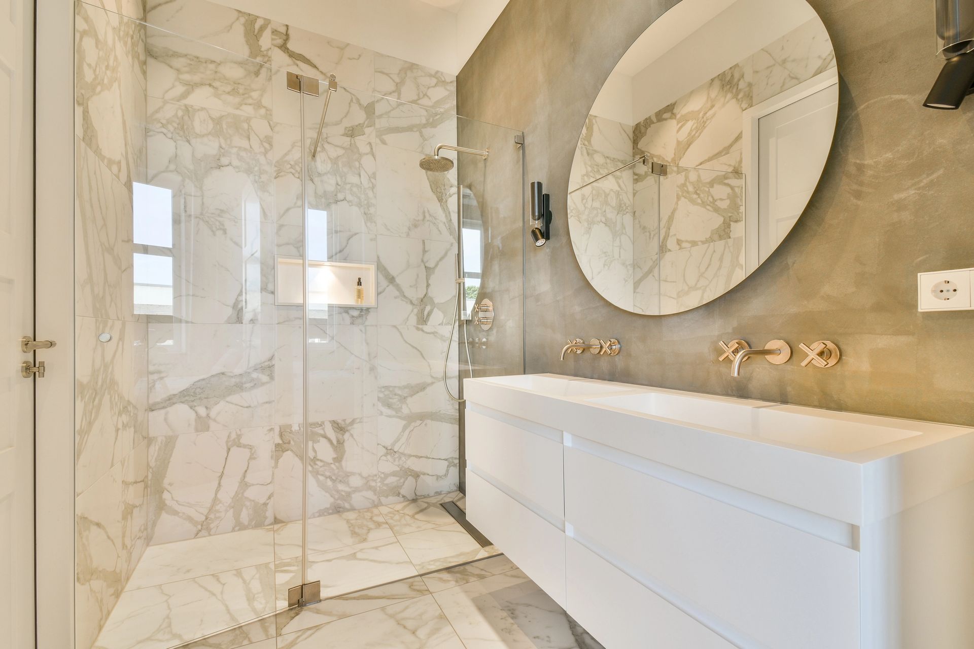 Baño moderno con ducha de mármol, tocador blanco y gran espejo ovalado.