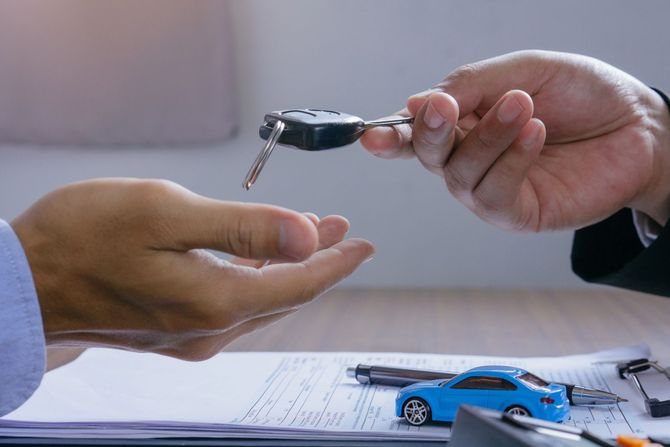 Due mani si scambiano le chiavi dell'auto sopra una scrivania, dove si trovano una macchinina blu giocattolo e dei documenti.