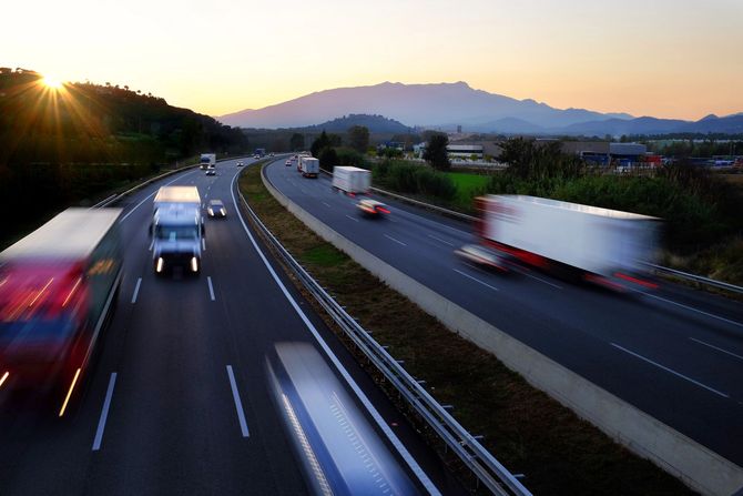 Vista al tramonto di un'autostrada trafficata con camion e auto sfocati su più corsie, montagne sullo sfondo.