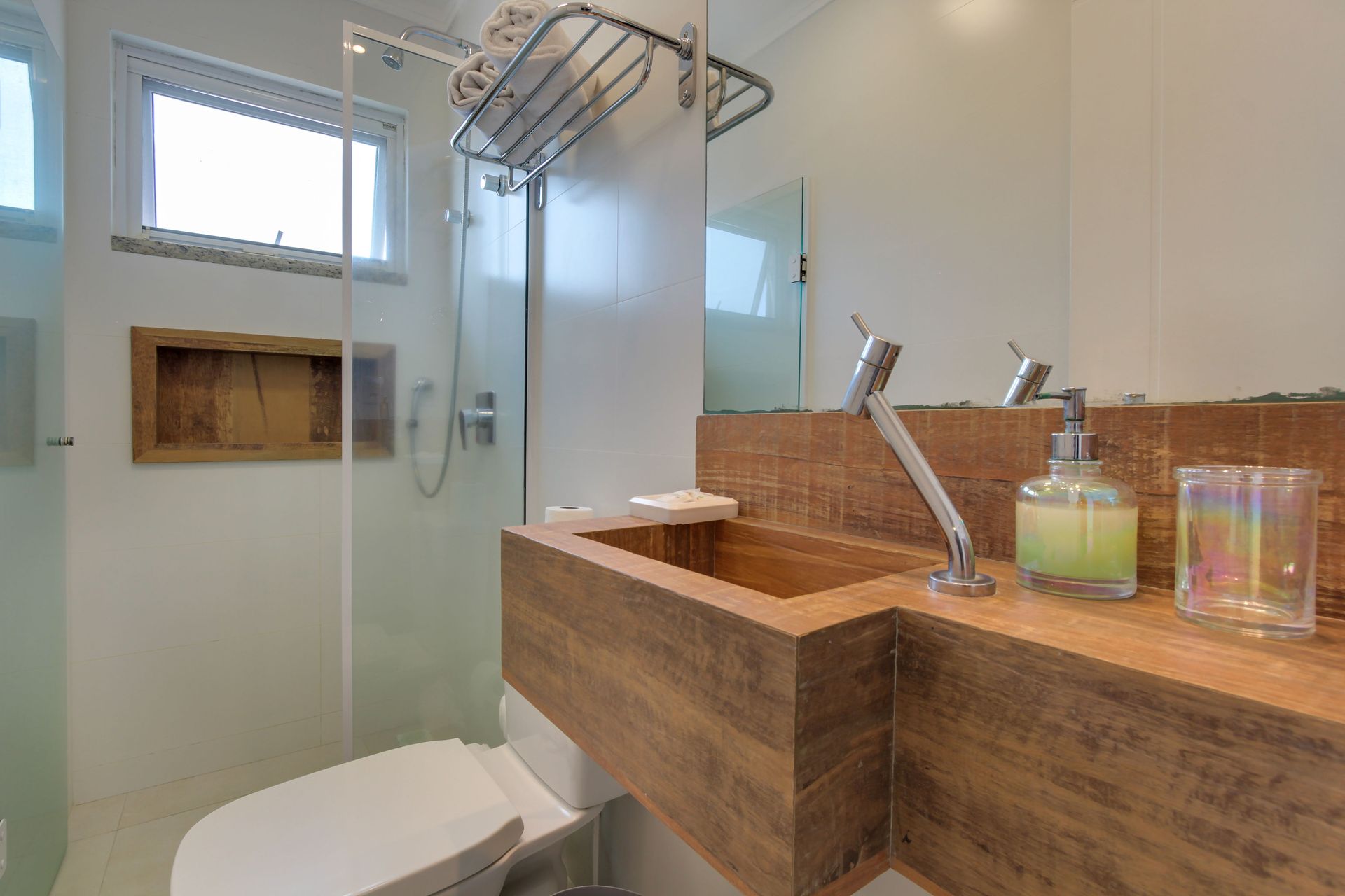 Banheiro moderno com pia de madeira, chuveiro e vaso sanitário. Paredes brancas e janela pequena.