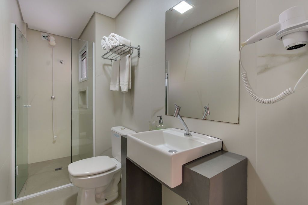 Banheiro com chuveiro, vaso sanitário, pia, espelho e porta-toalhas; tons neutros.