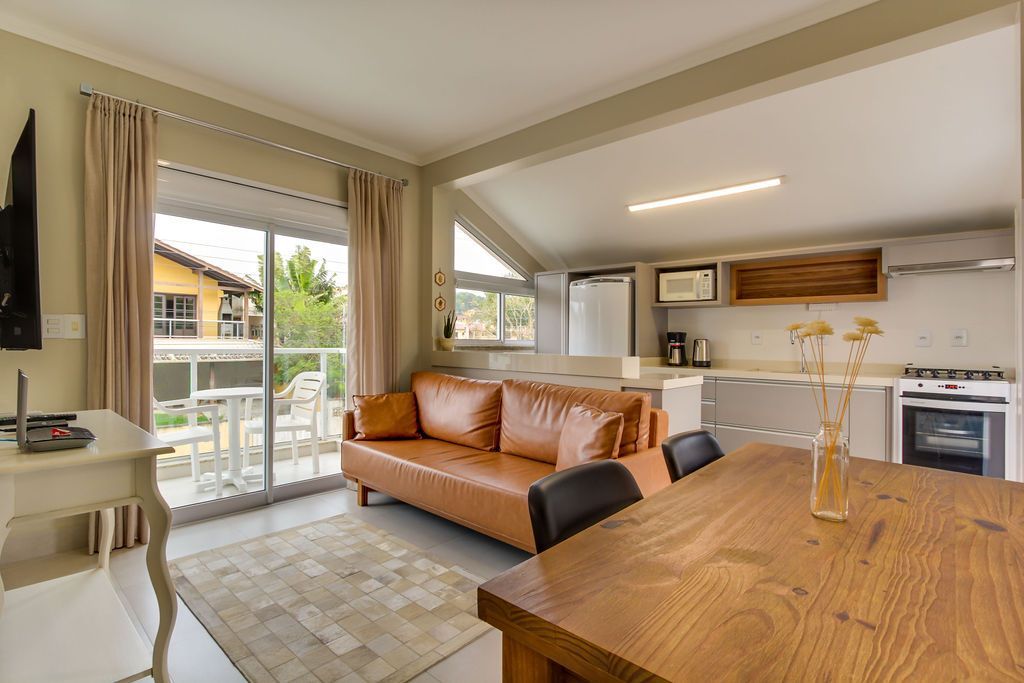 Sala de estar e cozinha com sofá marrom, mesa de madeira e porta de vidro deslizante para uma varanda.