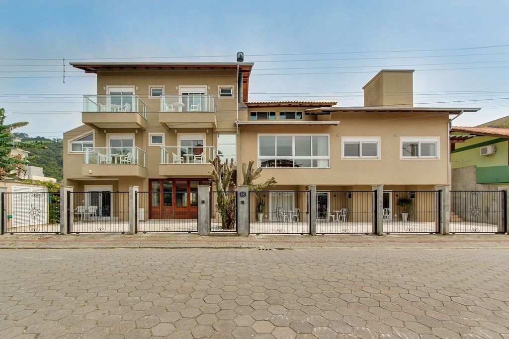 Prédio de vários andares em tom bege com varandas, rua de tijolos cinza e pátio fechado com portão.