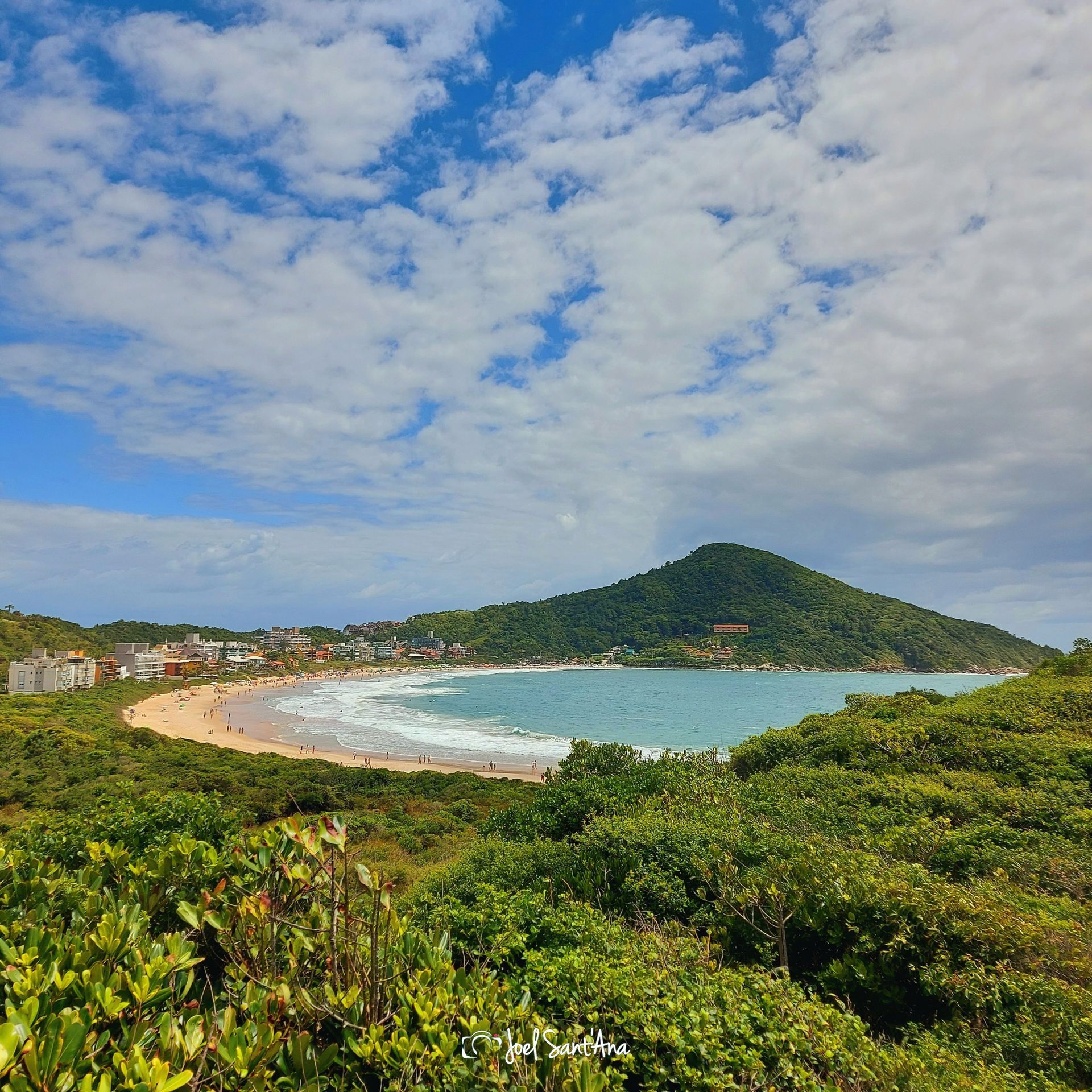 Praia com areia dourada, água azul e colina verde sob um céu parcialmente nublado.