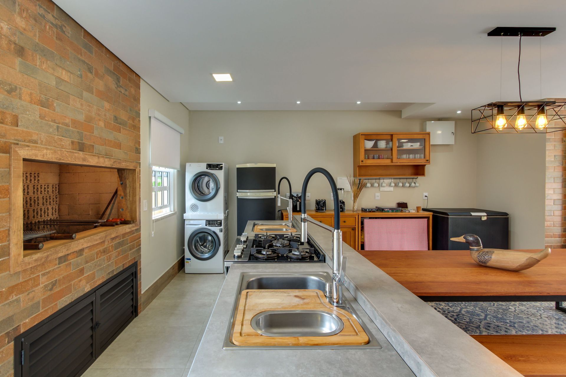 Cozinha externa com churrasqueira de tijolos, pia, fogão, eletrodomésticos e mesa de jantar.