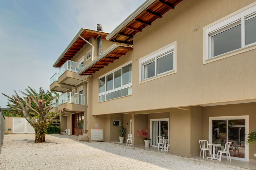 Casa de dois andares em tom bege, com varanda e janelas com moldura branca, situada em uma entrada de cascalho.