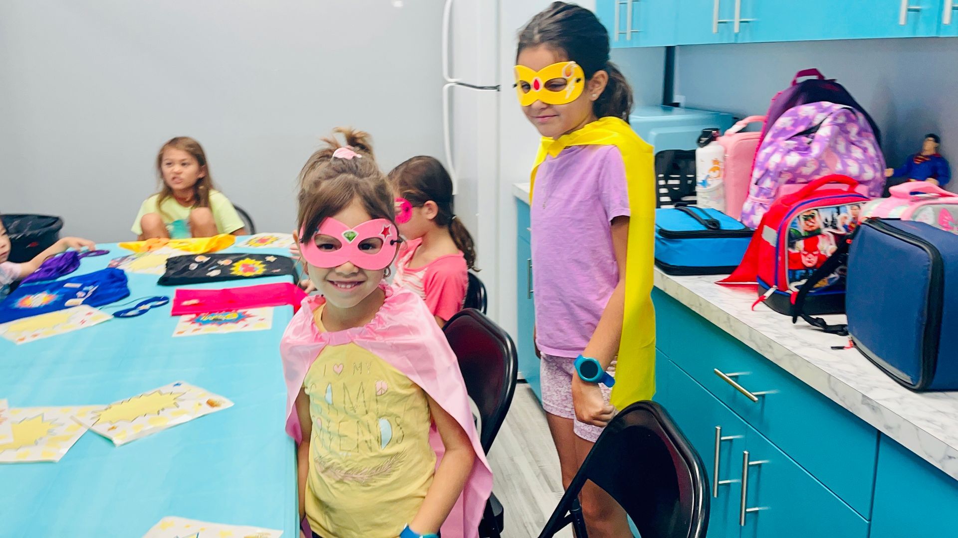Two little girls wearing masks and capes are standing next to each other in a kitchen.