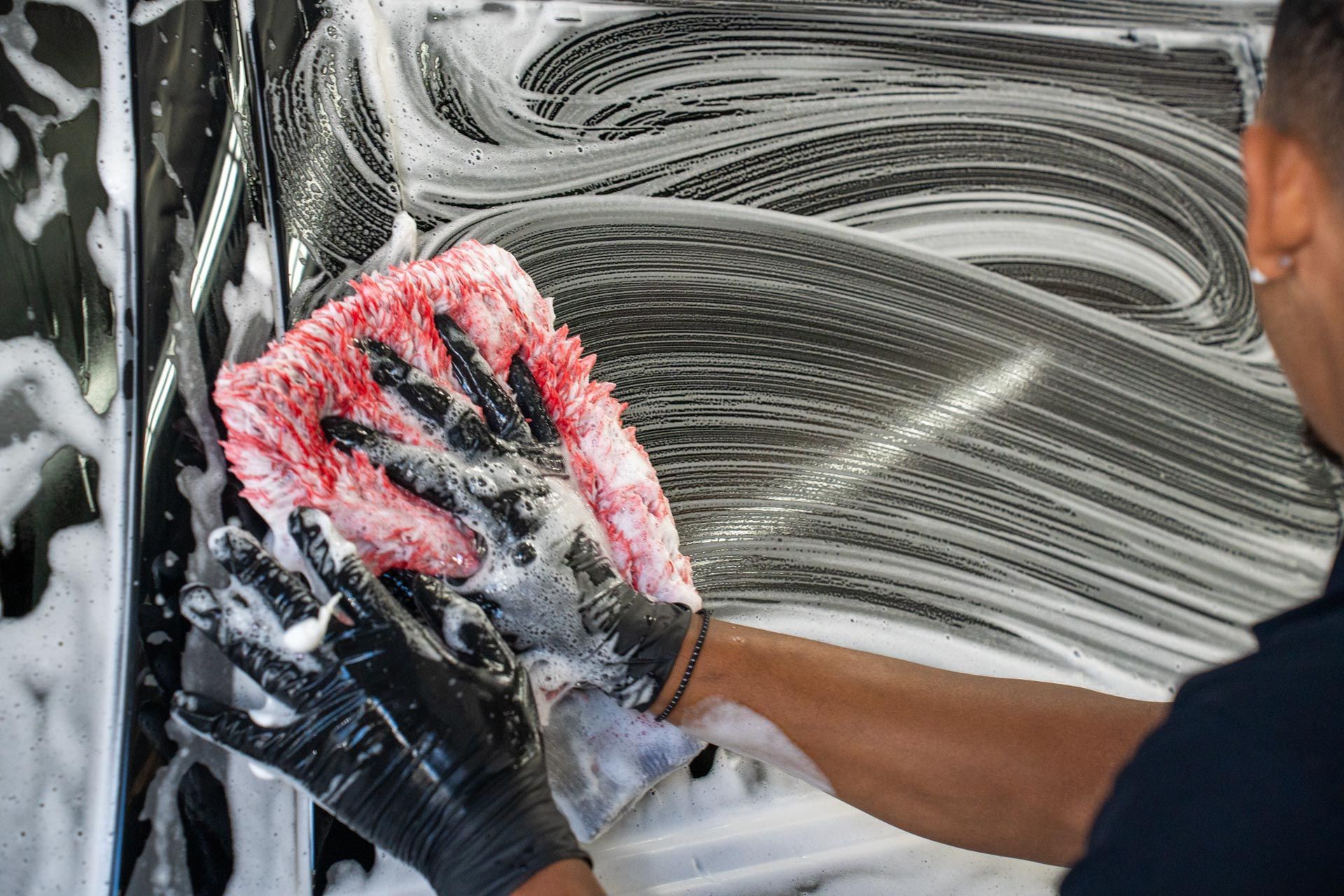Person washing a car with a red mitt and soapy foam, wearing black gloves.