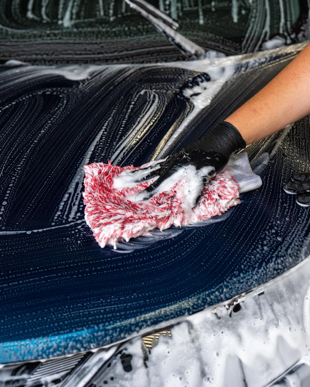 Person wearing black glove washing a car with soapy, red cloth.