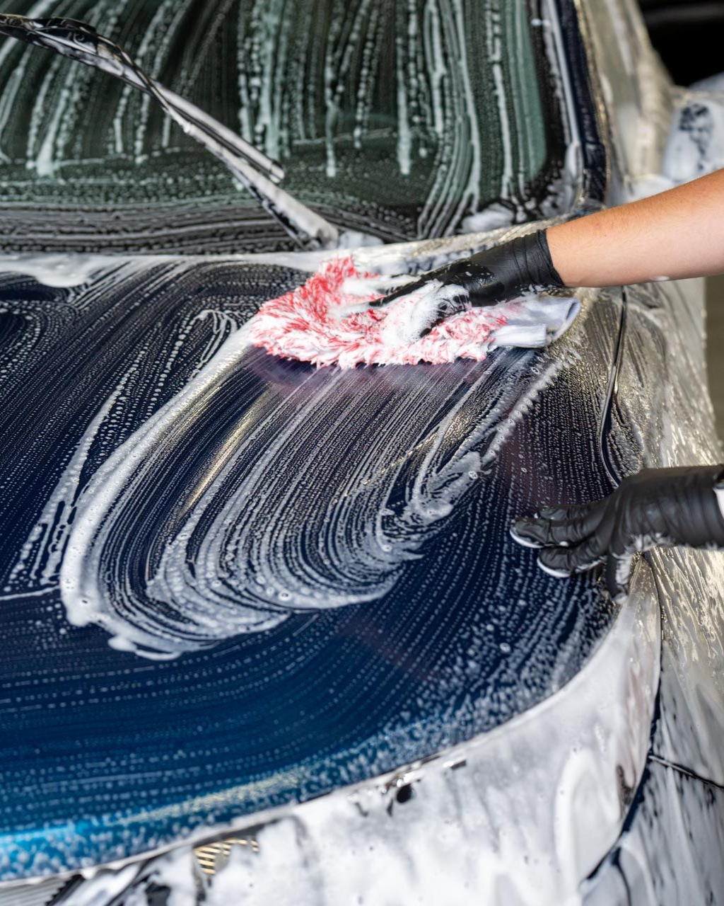 Car being washed with soapy foam; person in black gloves using a red wash mitt.