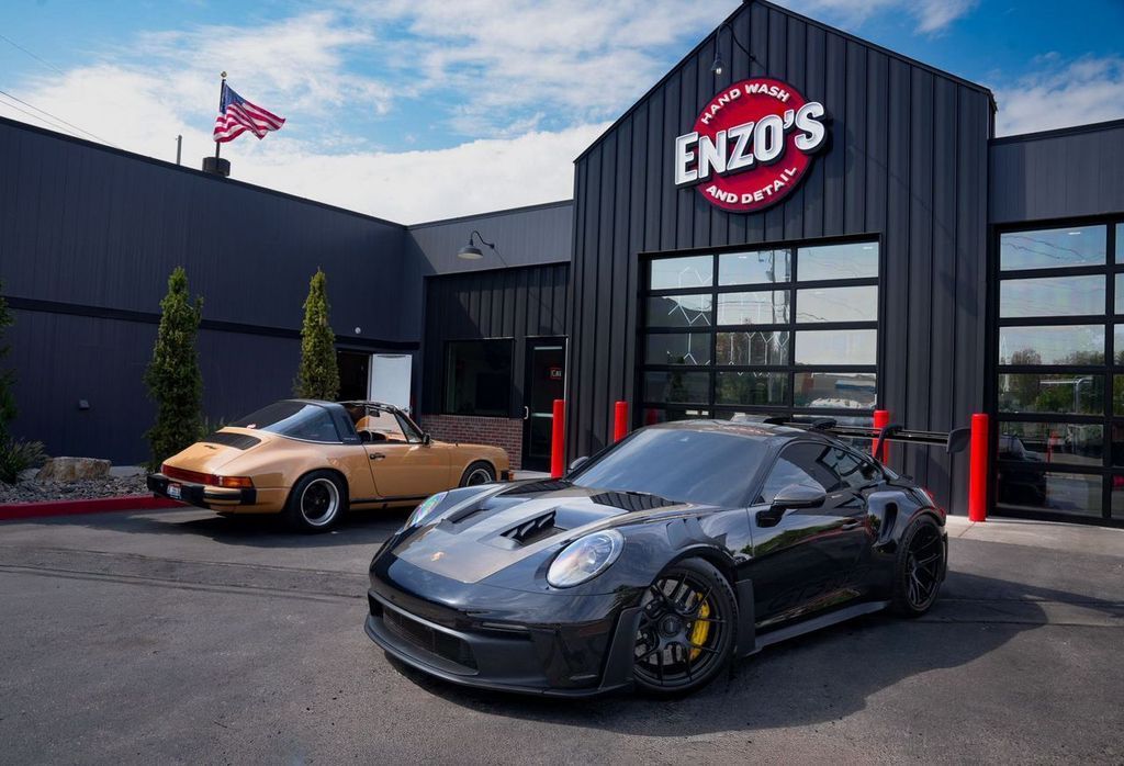 Man washing a black car, covered in suds, in a garage. Wearing a beanie and hoodie.