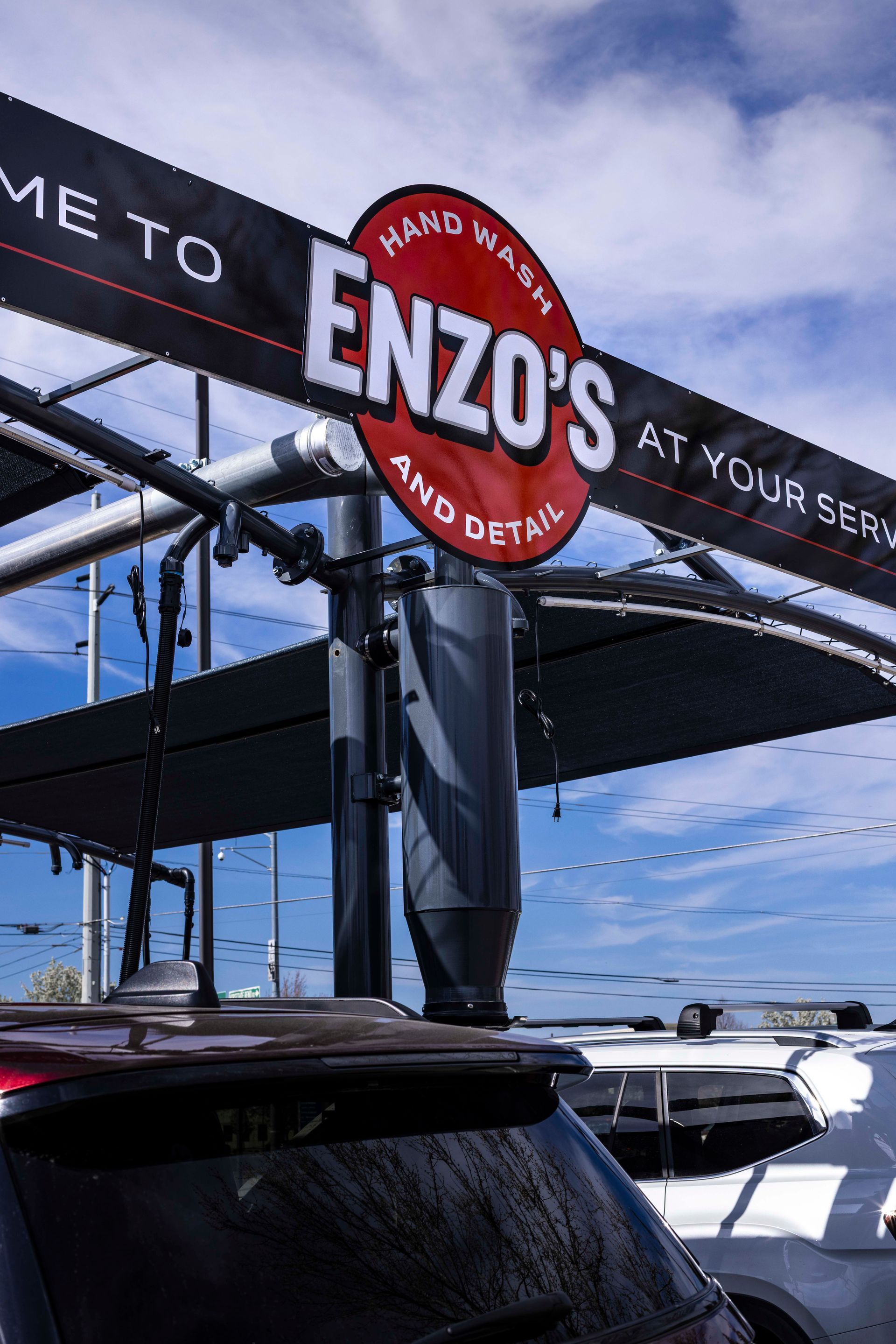 Sign for Enzo's Hand Wash and Detail. Red and white logo, black banner, car canopy, blue sky.