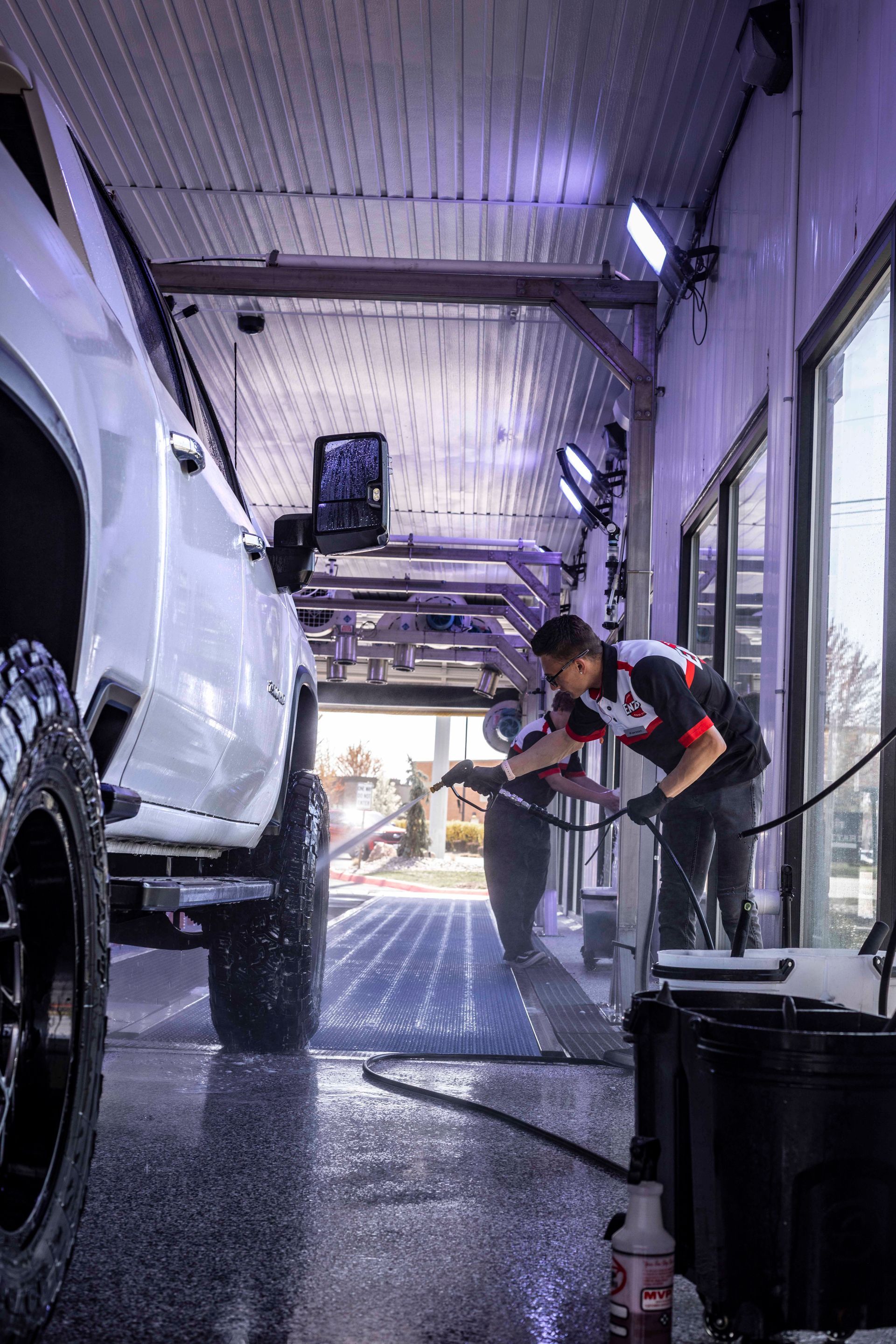Two men washing a white truck in a car wash bay. Soap and water are sprayed on the truck.