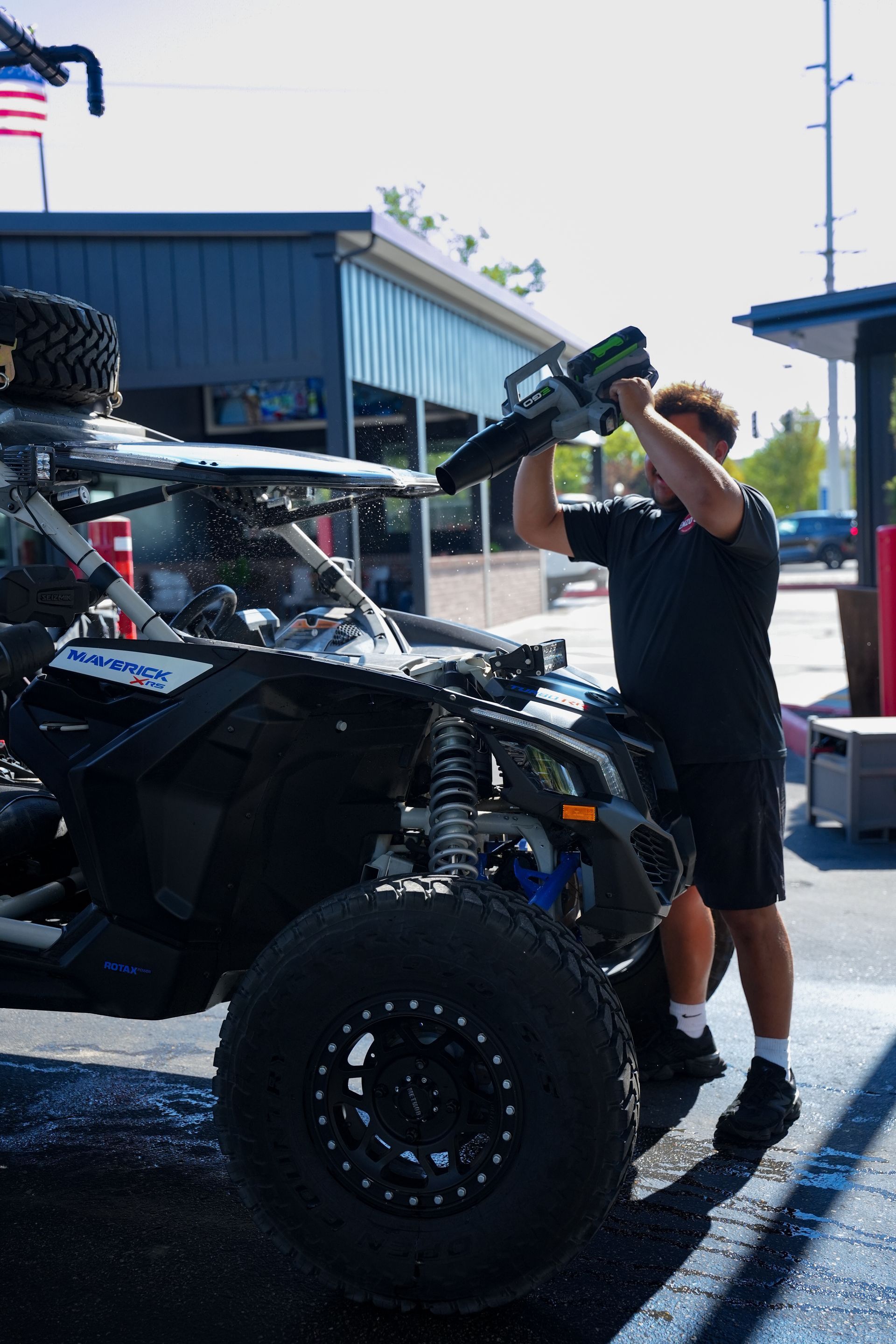Man washing a black and blue off-road vehicle with a pressure washer outside a building.