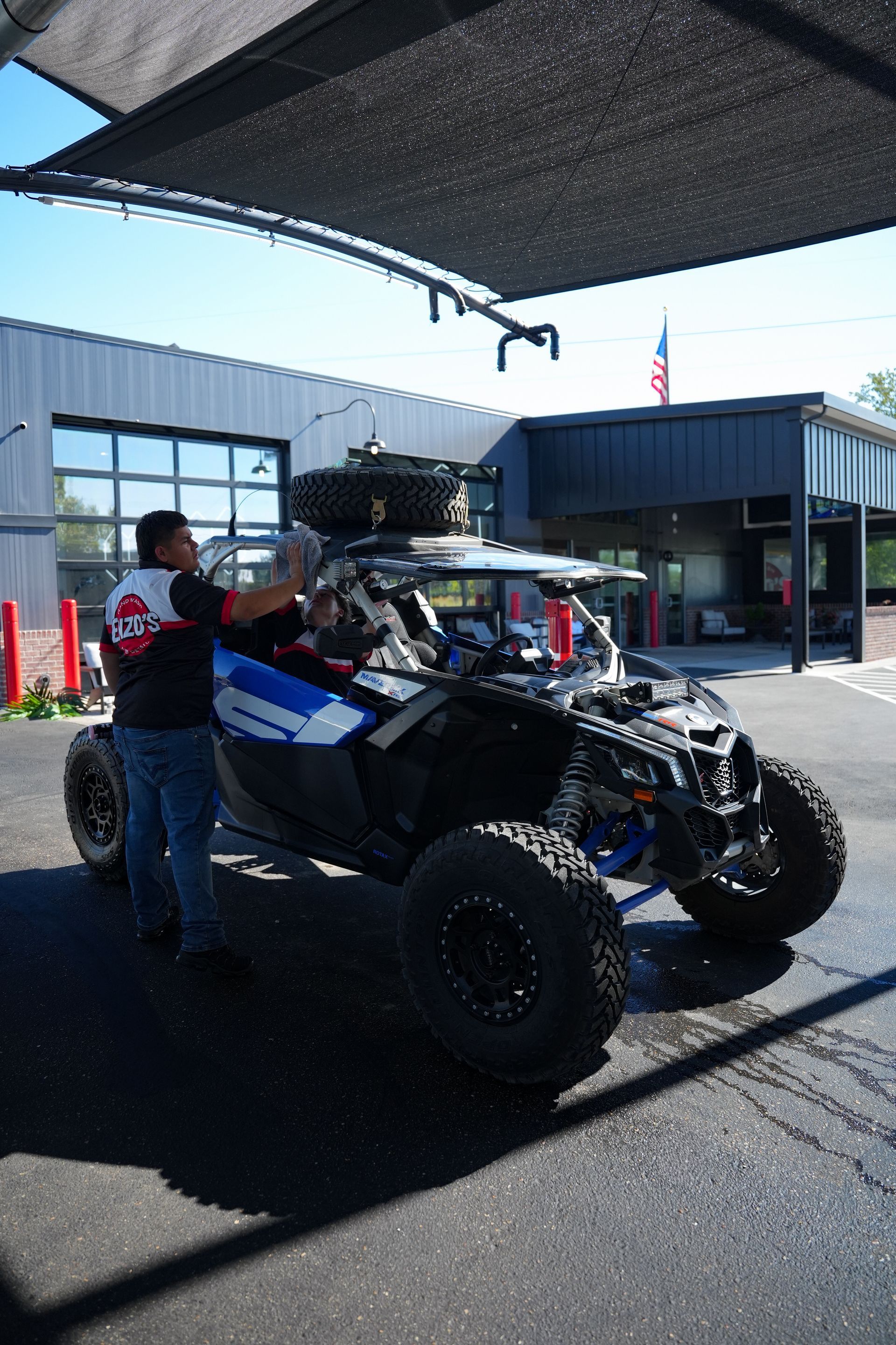 Man washing a black and blue off-road vehicle under a shaded structure, another tire on top. Building background.
