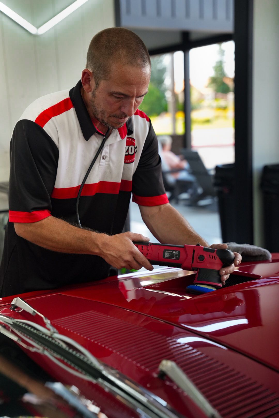 Man polishing a red car with a buffer in a workshop. He wears a red and black shirt.