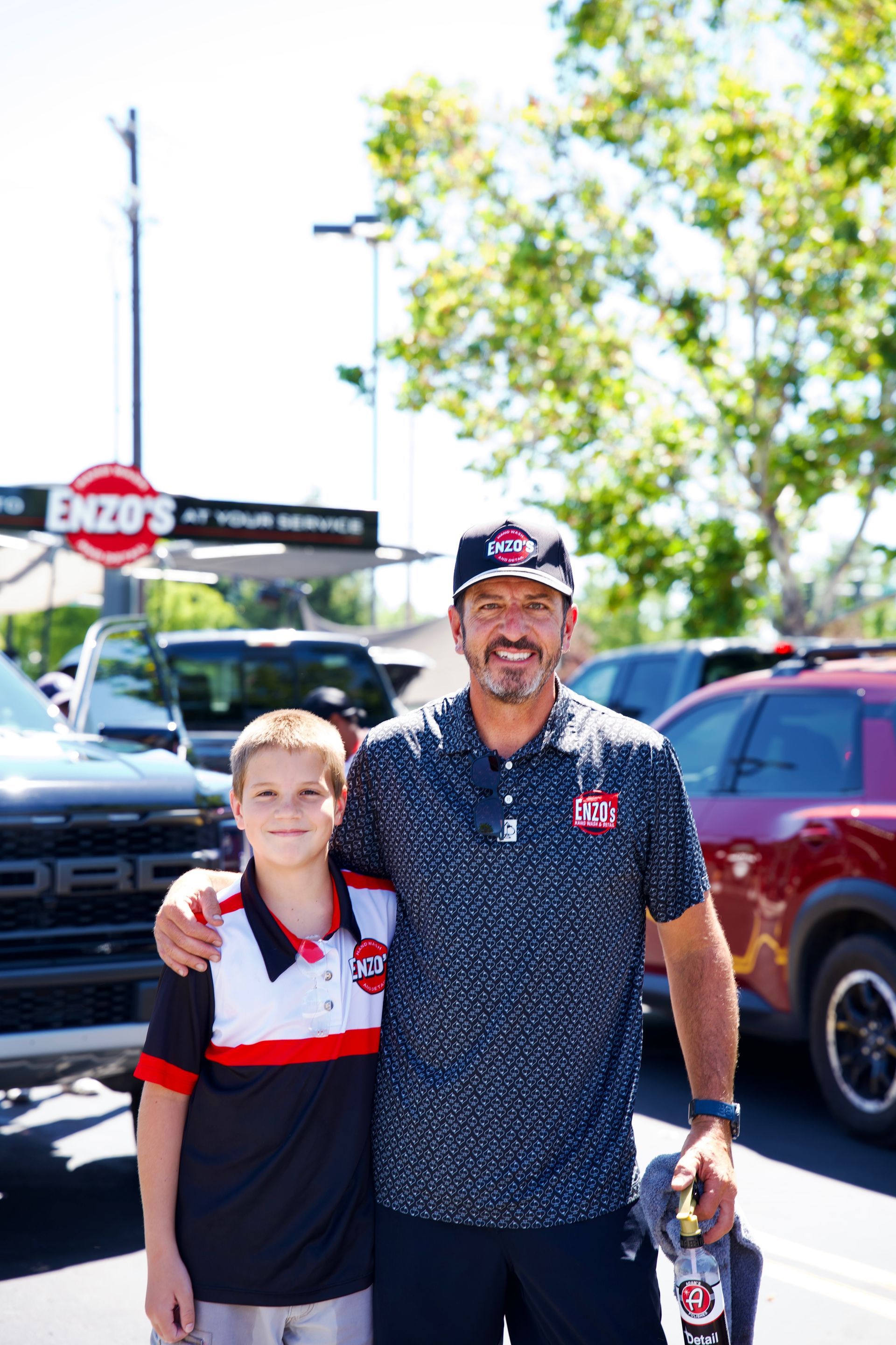 Man and boy stand outside Enzo's, man's arm around boy. Cars and sign in background. Sunny.