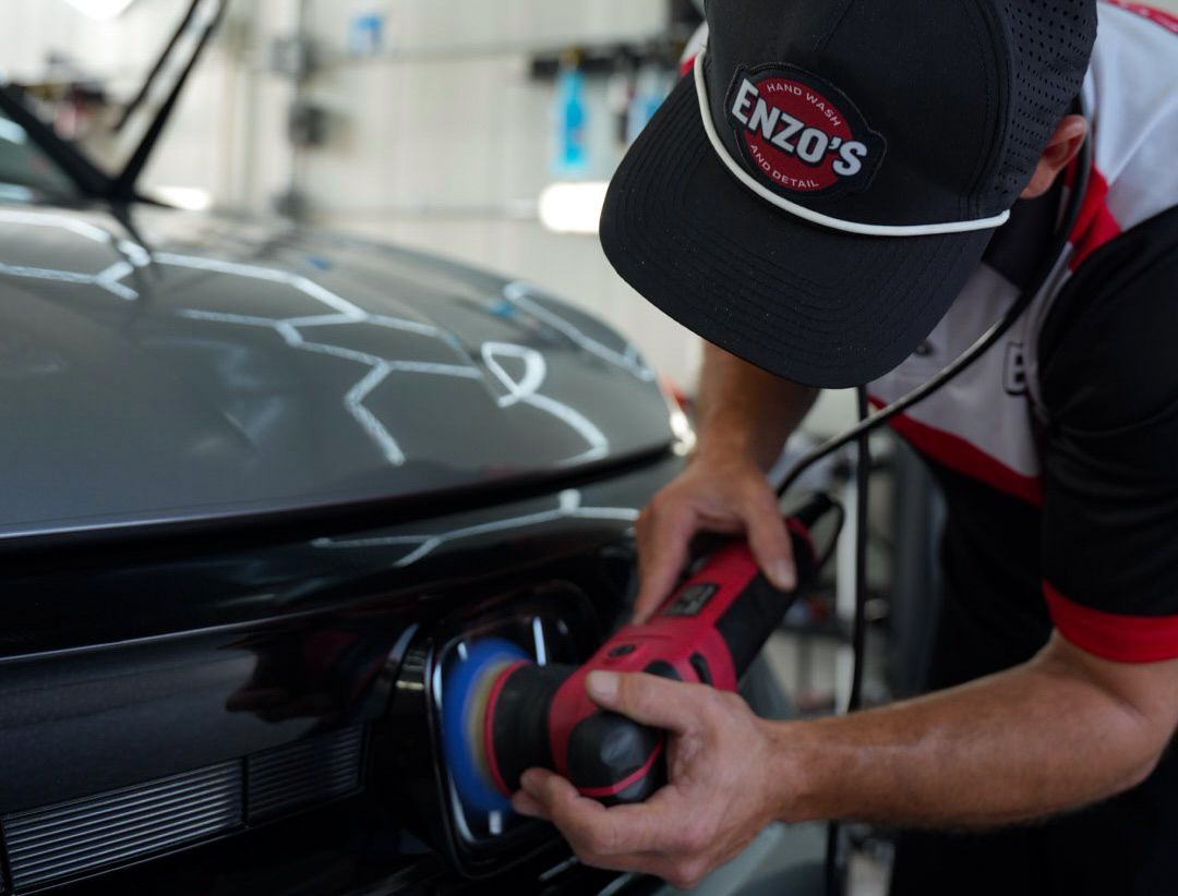 Person polishing car emblem with a red and black tool, wearing a hat that reads "Enzo's".