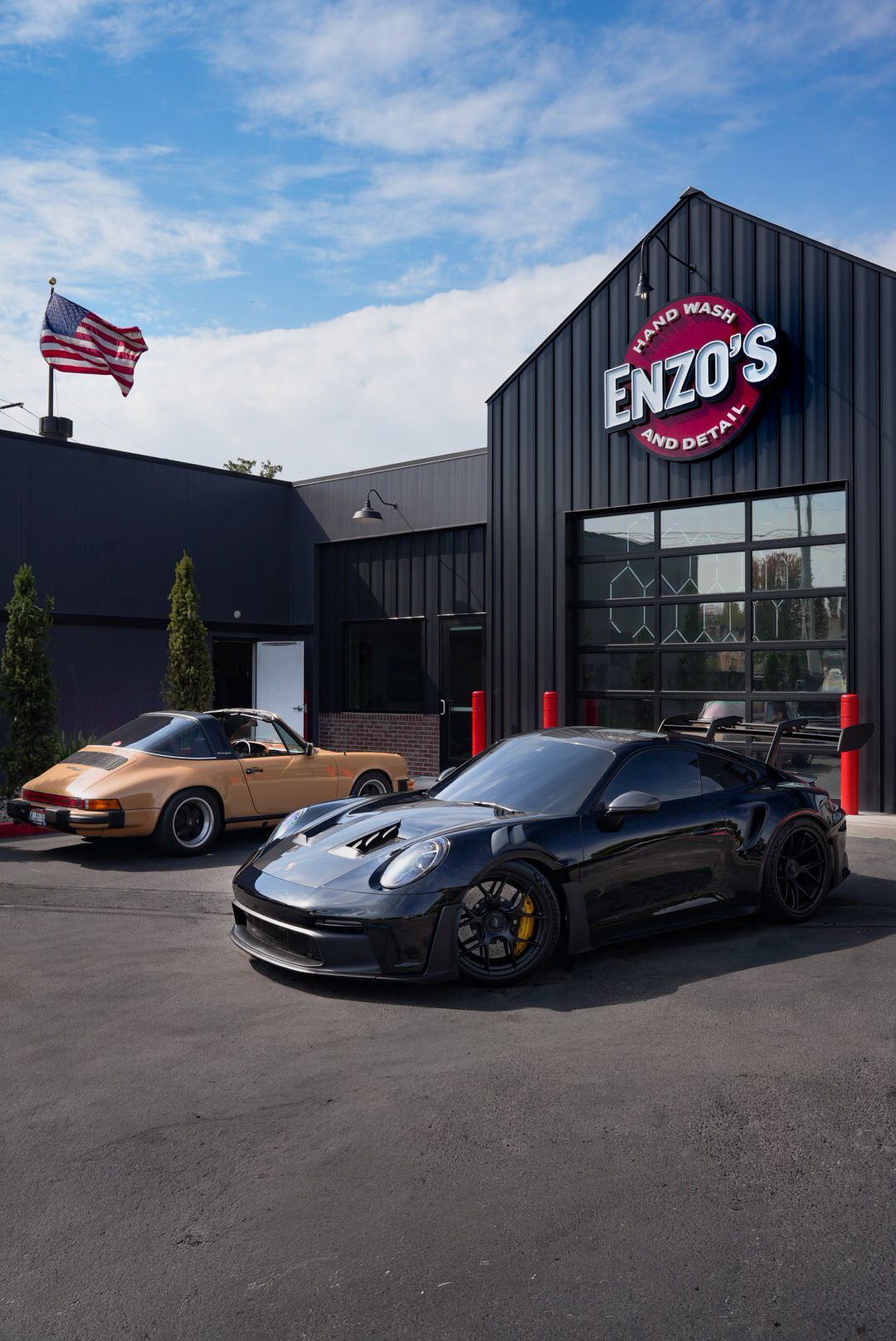 Two Porsche sports cars parked in front of Enzo's Auto Care. Black and gold exterior, blue sky, American flag.