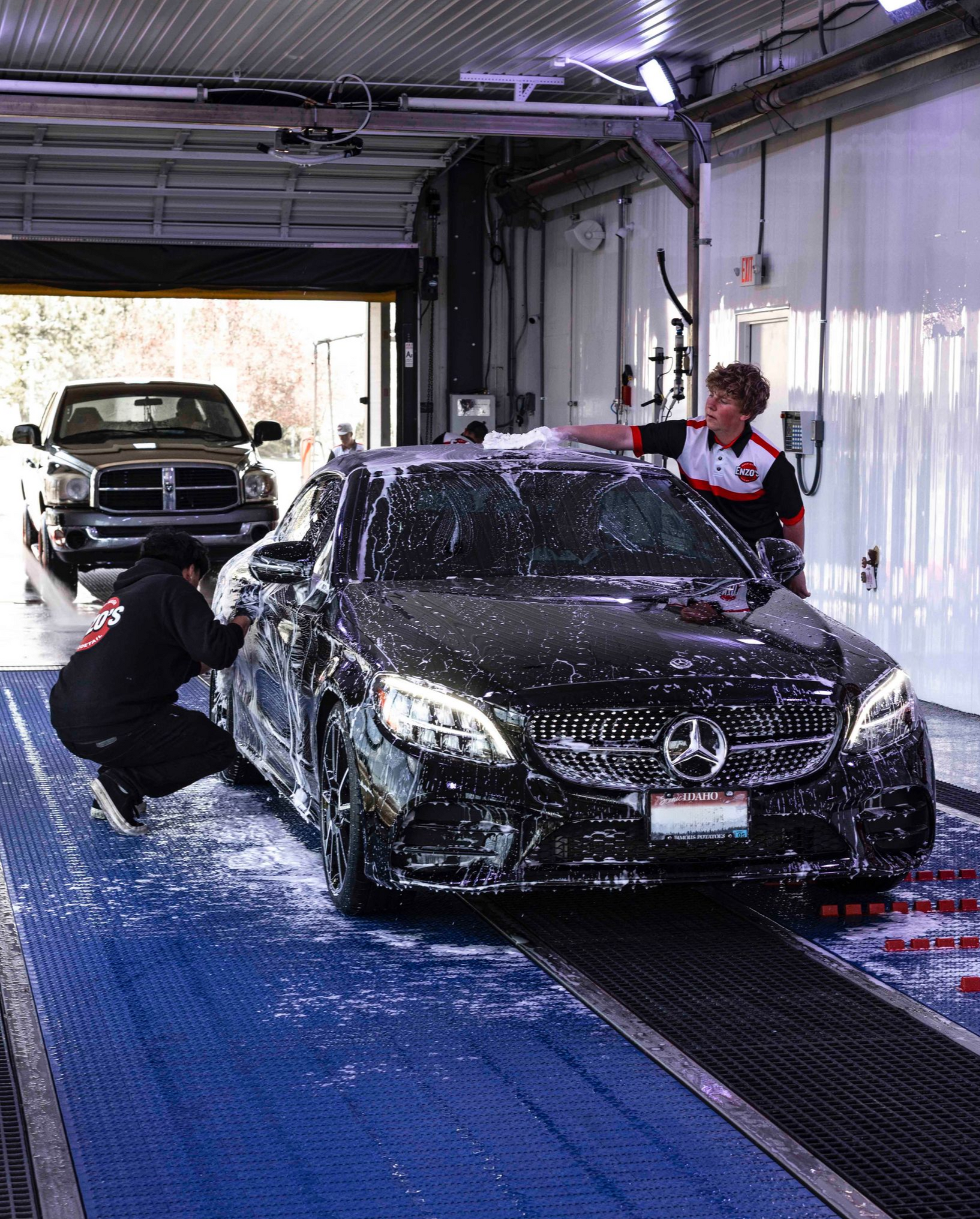 Two people washing a black car at a car wash. The car is covered in foam. A truck waits behind.