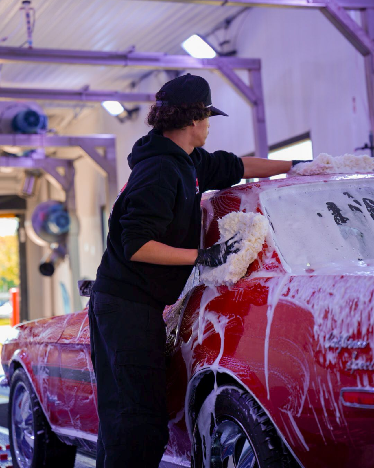 Person washing a red classic convertible car with a sponge at a car wash.