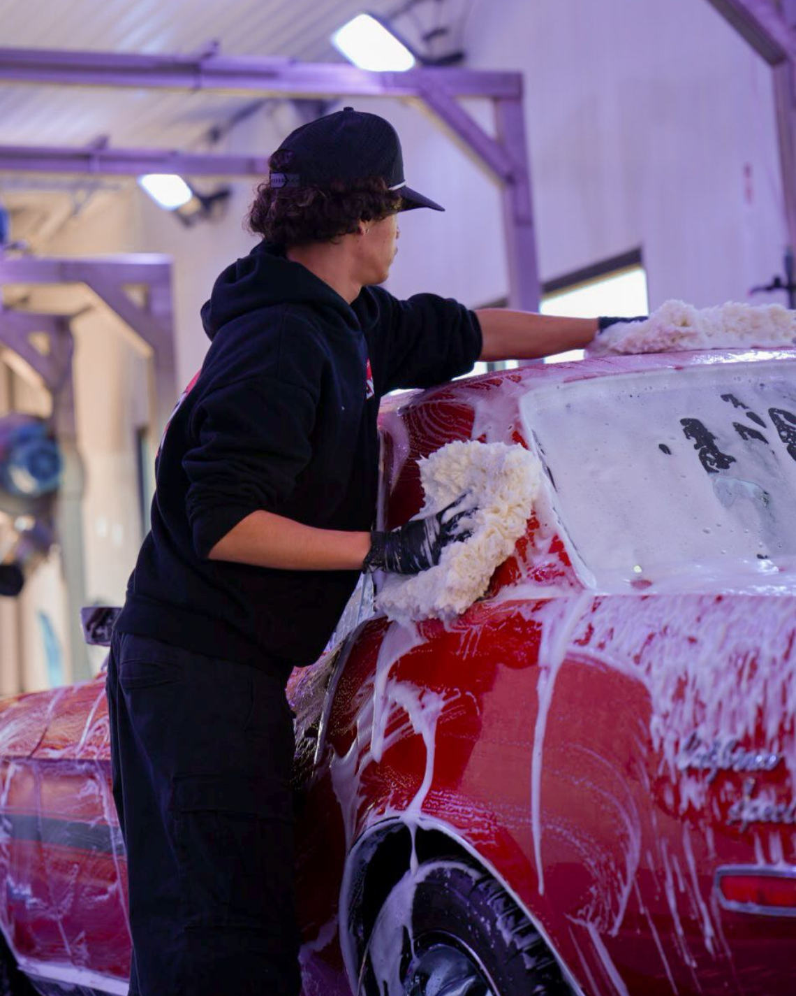 Person washing a red car with soap in a garage, wearing a black hat and hoodie