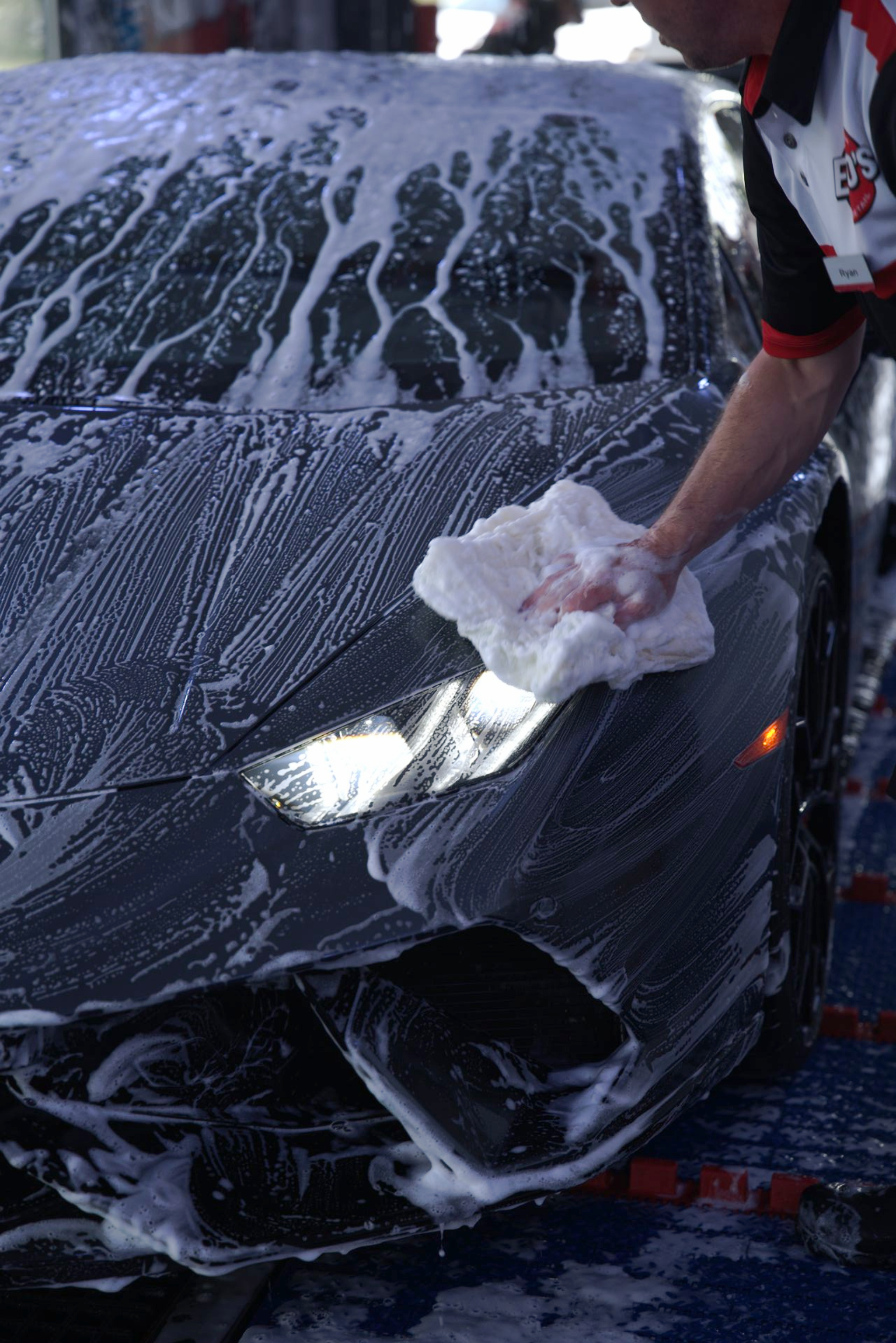 Car being washed with soapy foam; person in black gloves using a red wash mitt.