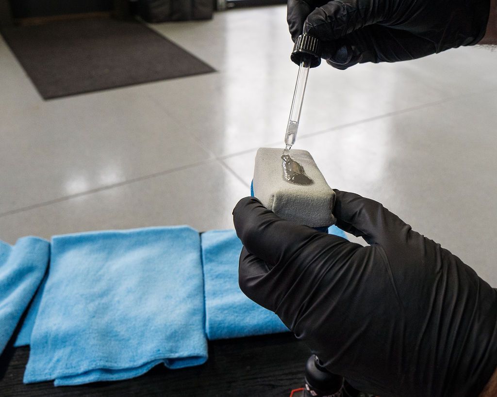 Gloved hands applying a liquid to a gray applicator pad. Blue microfiber towels are nearby on a table.