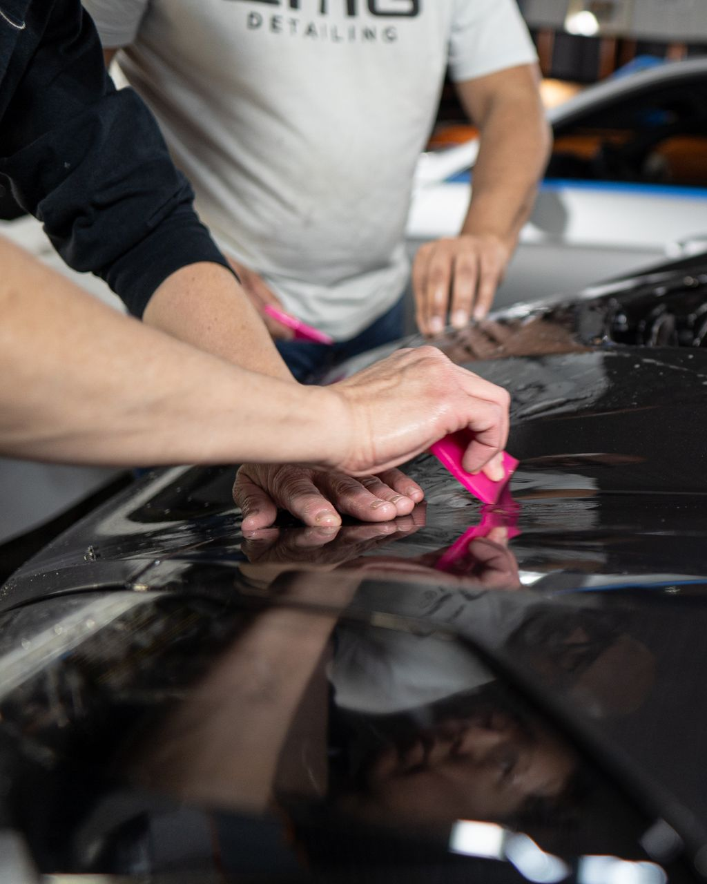 Two people applying protective film to a black car hood with pink squeegees in a garage.