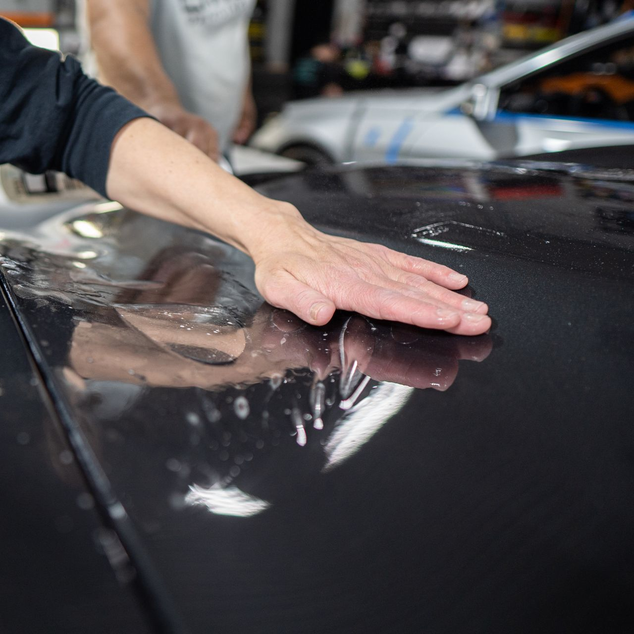 Person smoothing a clear film onto a black car hood in a garage.